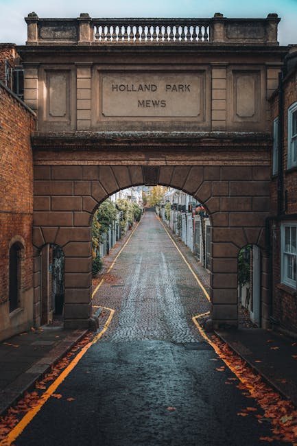 Photograph of a narrow cobbled street viewed through a large stone archway marking the entrance to Holland Park Mews, with the inscription 'Holland Park Mews' engraved on the stone above the arch. On either side of the street, there are brick and stone buildings with small windows, some of which have decorative frames. The street has a slight incline and is lined with parked cars and greenery, with trees visible in the distance. The scene captures a quiet, residential area possibly during early morning or late afternoon, with soft natural lighting. This setting may be used in the context of house removals or moving services, such as those offered by [COMPANY_NAME], illustrating the typical environment for furniture transport or packing and moving logistics in Holland Park.