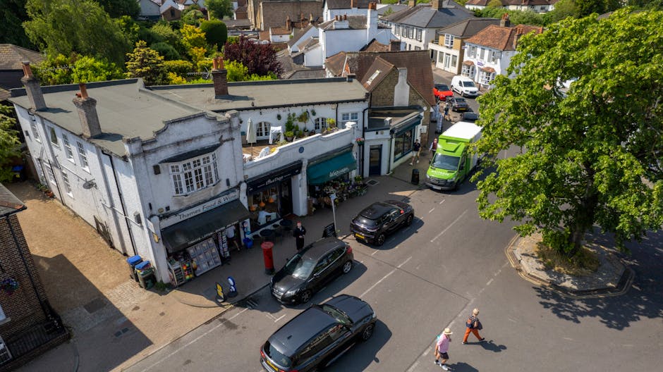 An aerial view of a street in Holland Park showing a two-storey building with a white facade and black awnings on the ground floor, which hosts small shops. Several cars, including a black hatchback, a black estate, and a green van, are parked along the street, with the green van positioned near the building entrance. The pavement in front of the shops features a red post box, a blue recycling bin, and a few pedestrians, some of whom are walking with shopping bags or wearing coats. To the right, a large green tree provides shade for part of the street, and additional residential houses with tiled roofs are visible beyond. The scene depicts an active urban environment, potentially involved in a house or office relocation process with transport vehicles and pedestrians engaged in their daily activities, exemplifying the context of removals and moving logistics supported by [COMPANY_NAME].