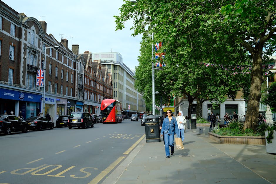 Photograph of a narrow cobbled street viewed through a large stone archway marking the entrance to Holland Park Mews, with the inscription 'Holland Park Mews' engraved on the stone above the arch. On either side of the street, there are brick and stone buildings with small windows, some of which have decorative frames. The street has a slight incline and is lined with parked cars and greenery, with trees visible in the distance. The scene captures a quiet, residential area possibly during early morning or late afternoon, with soft natural lighting. This setting may be used in the context of house removals or moving services, such as those offered by [COMPANY_NAME], illustrating the typical environment for furniture transport or packing and moving logistics in Holland Park.