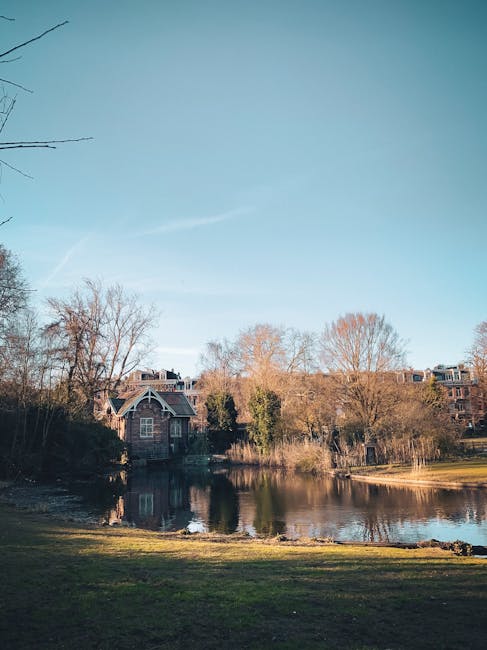 A tranquil lakeside scene with calm water reflecting the surrounding trees and buildings, including a large brick house with a gabled roof and multiple windows situated near the shoreline. In the foreground, there is a grassy area with some low shrubbery, while the background features leafless trees indicating late autumn or early winter. The sky above is clear with a few wispy clouds. This serene setting is typical for a residential area where house removals or furniture transport might occur, with the house potentially being part of a home relocation or move managed by [COMPANY_NAME], utilizing packing and loading procedures suitable for outdoor and lakeside environments.
