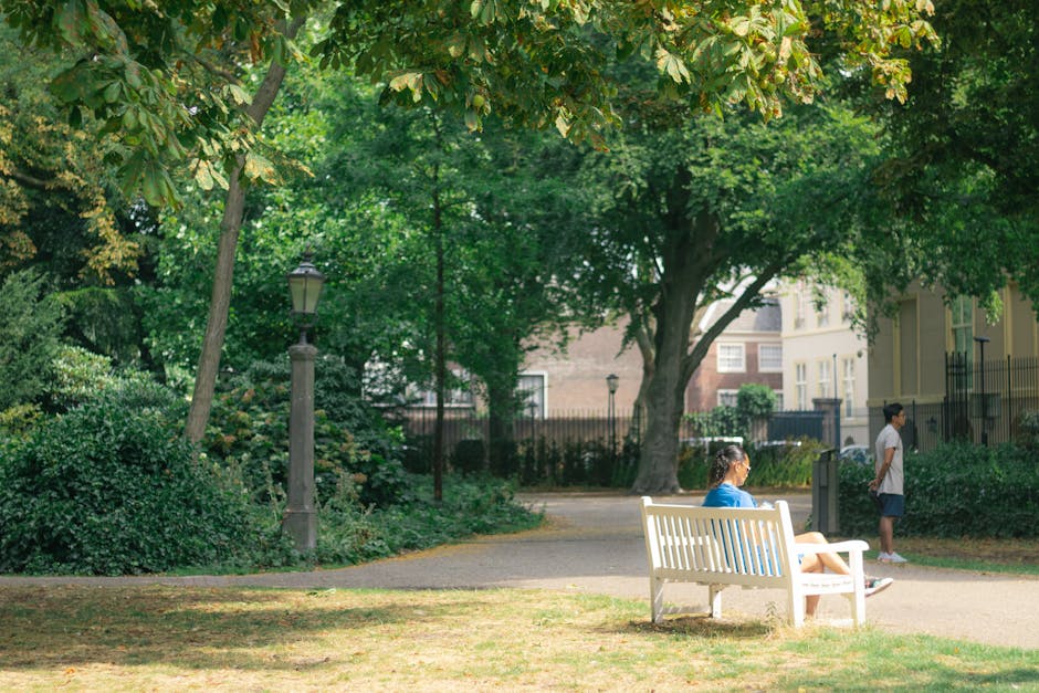 A woman in a blue shirt and shorts is seated on a white park bench in a lush, green urban park, surrounded by trees and shrubbery. She appears to be resting or waiting, with her legs crossed and looking towards the left. Nearby, a man in a white shirt and dark shorts is standing on the paved pathway, possibly observing the surroundings. The park features a lamppost, a mix of mature trees providing shade, and residential buildings with traditional architecture in the background. The scene is well-lit by natural daylight, emphasizing the calm and tidy environment suitable for casual walks or resting during home relocations. This setting captures the tranquil atmosphere often encountered during moving or packing activities, with the outdoor space arranged to support a smooth furniture transport and loading process, highlighting the importance of outdoor access for house removals and relocation services provided by [COMPANY_NAME].