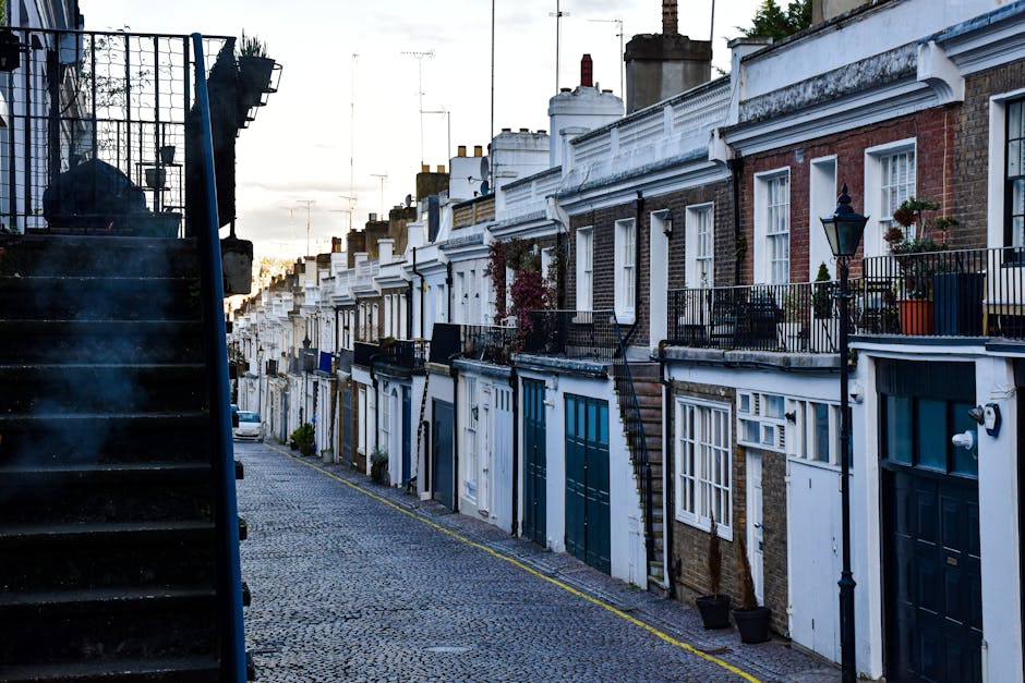 A row of traditional Victorian terraced houses in Holland Park, London, viewed from the cobbled pavement sidewalk. The buildings feature brick facades, white decorative cornices, and small front gardens with potted plants and outdoor lighting fixtures. A black metal staircase ascends beside one of the houses, leading to the upper floor. Several houses have black wrought iron balconies and railings, some with flower pots or small plants. The street is narrow, with a slight incline, and the sky above is partly cloudy with the setting sun casting a warm light over the scene. This environment is typical of home relocation and furniture transport processes, with a focus on the residential street setting where removals services by [COMPANY_NAME] may be provided as part of house removals or moving logistics.