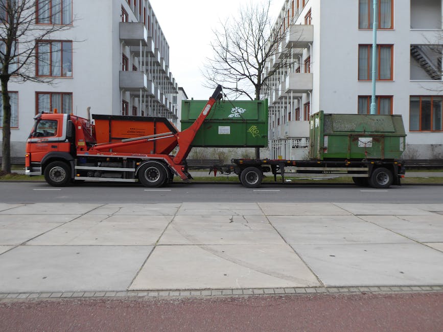 A white and dark blue garbage disposal truck, equipped with hydraulic lifting mechanisms at the rear, is parked on a street in front of a multi-storey brick building with numerous rectangular windows.
