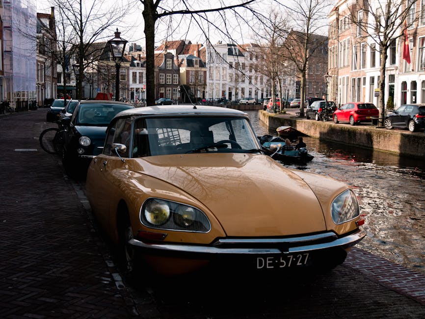 A vintage yellow sports car with a soft black roof, parked on a cobbled street next to a canal in a residential area. The car's front license plate reads 'DE 57-27.' Behind it, there is a black vehicle with a bicycle mounted on the rear, both situated alongside other parked cars along the street. The canal, visible on the right, has a brick embankment and a few small boats moored nearby. In the background, traditional European-style townhouses with large windows, gabled roofs, and facades featuring various shades of brick and plaster are lined along the canal. Leafless trees are present along the street, indicating a winter or early spring season, with natural daylight illuminating the scene. The environment suggests a quiet neighbourhood suitable for home relocations and furniture transport, with [COMPANY_NAME] potentially servicing such moving logistics in this area.